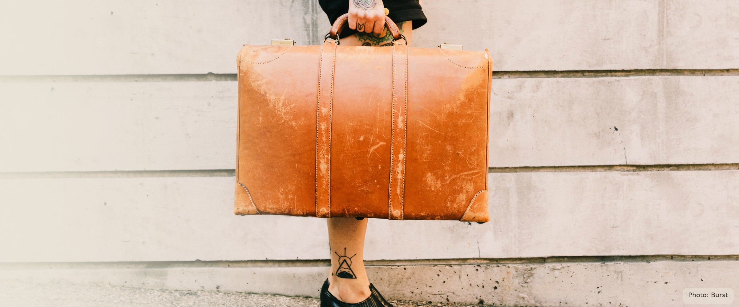 A young woman's hand carries a worn vintage suitcase against a plain limestone wall - starting adult life with what she can carry.