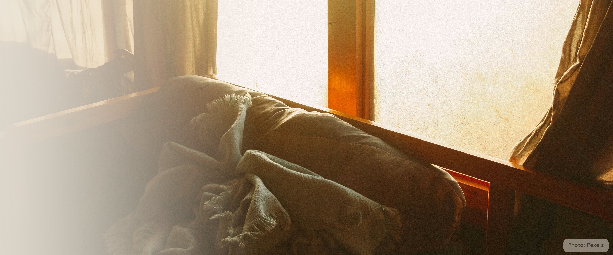 A folded blanket rests on a worn sofa cushion, warm window light behind - the hidden kind of homelessness.